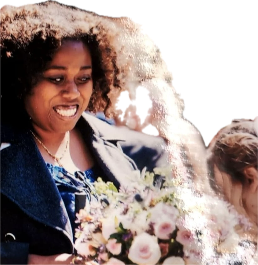 Lily, a Black woman with natural, curly brown hair which frames her face is smiling broadly She is looking down towards a large, varied bouquet of flowers she holds, which features soft pink, cream, white, and purple blooms with green foliage. She is wearing a grey pea coat over a blue patterned top and a gold chain necklace.